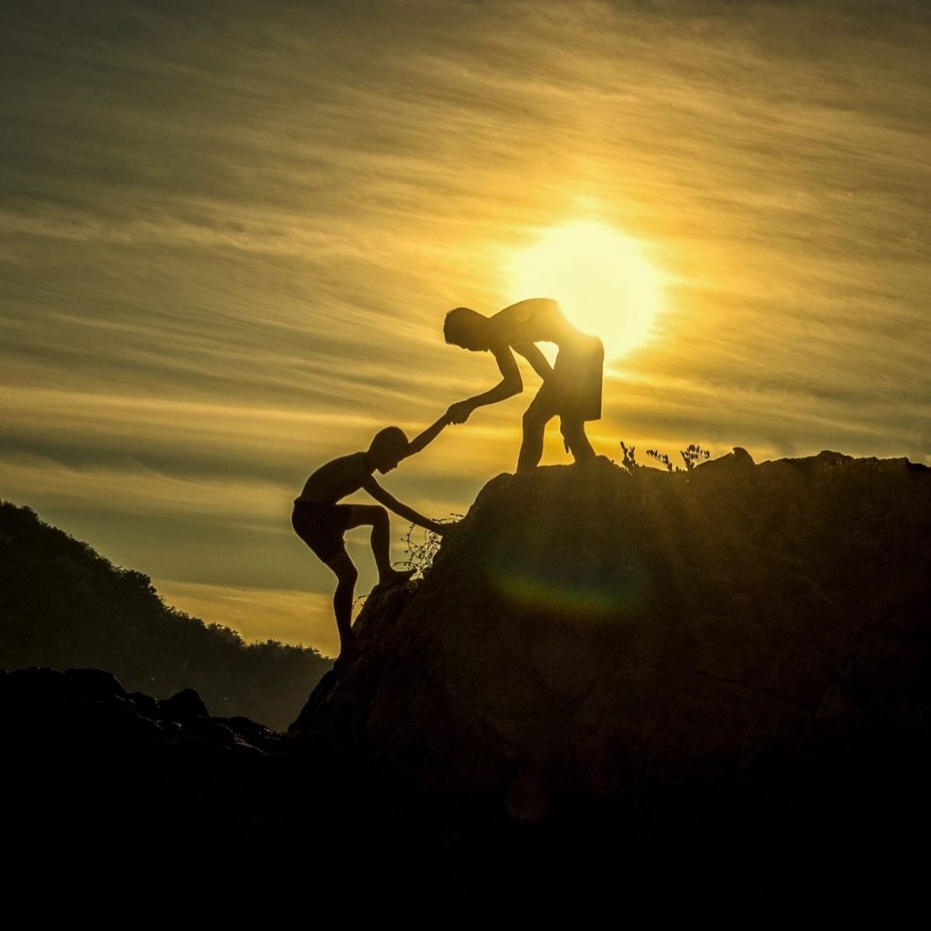 One person helps another climb a mountain with a sunset in the background.